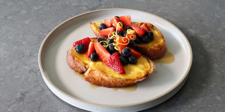 A plate of golden French toast topped with fresh strawberries, blueberries, a drizzle of syrup, and a garnish of lemon zest, served on a light gray plate on a dark countertop.